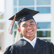 A young man smiling into the camera, donning his graduation cap and gown in front of a glass block facade