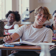 A young man, leaning back, smiling into the camera, sitting at his desk with a book open in a classroom