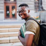 A young man in glasses smiling into the camera as he walks away towards the steps of his school, books in tow