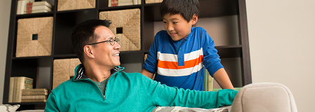 Father sitting on couch smiles at his son, who is standing up.