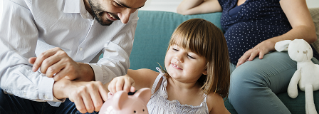 Dad helps young daughter place some coins in her piggy bank as pregnant mom watched from the couch.