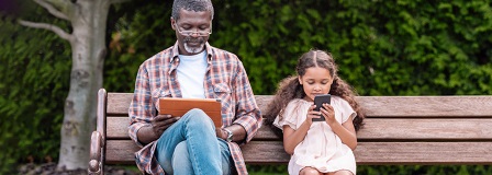 Grandfather and granddaughter looking at electronics while sitting on park bench wh