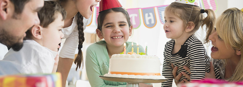 Smiling girl standing behind a birthday cake while family is singing to her