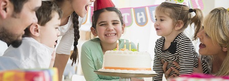 Smiling girl standing behind a birthday cake while family is singing to her