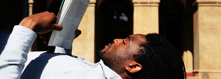 Man reading a book while lying on the the ground in front of college building