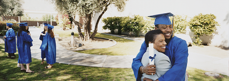 A man, dressed in graduation cap and gown, hugs his mom while holding his diploma.